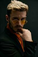 blond man with glasses and black jacket orange sweater portrait dark background photo