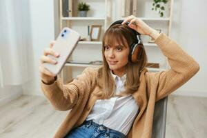 Cheerful young blonde lady in warm sweater in headphones listen fav songs doing selfie video call sitting in armchair at modern home interior. Music time Relaxing Cool playlist Concept. Copy space photo