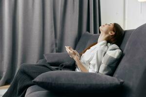 mujer con móvil teléfono en sofá mirando arriba y cortinas en el antecedentes foto