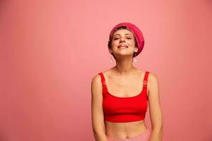 Young athletic woman with a short haircut and purple hair in a red top and a pink hat with an athletic figure smiles and grimaces looking at the camera on a pink background photo