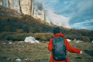 a woman in a red jacket with a backpack and in a warm hat walks on the grass in the fall in the mountains photo