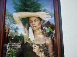 woman holds hand on forehead in medical mask at window photo