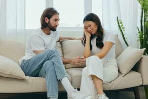 A man and a woman of different races sit on the couch in a room at home and talk about their problems to each other. A stress-free lifestyle of family quarrels with psychological support photo