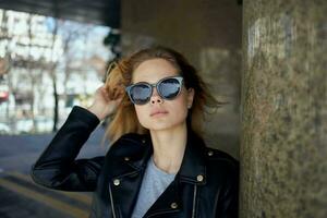 un mujer soportes debajo un puente en el calle vistiendo Gafas de sol y un cuero chaqueta foto