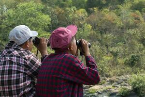 Asian boys sitting, holding binoculars and looking up to watch the birds which flying on sky and sitting on tree branches in local national park to study birds, nature, environment and landscape. photo