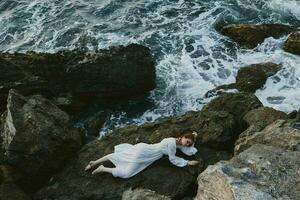 atractivo mujer con largo pelo en un blanco vestir acostado en un Roca en un blanco vestir vacaciones concepto foto