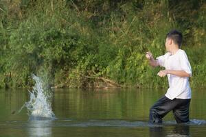 asiático chico en blanco camiseta es gasto su tiempo libre por buceo, nadar, lanzamiento rocas y atrapando pescado en el río felizmente, pasatiempo y felicidad de niños concepto, en movimiento. foto