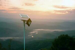 mini fotovoltaica o solar célula panel instalado en metal polo con hd Luz de inundación a Tienda y utilizar el poder desde el luz de sol a noche alrededor casa en el montaña, borroso puesta de sol montaña antecedentes. foto