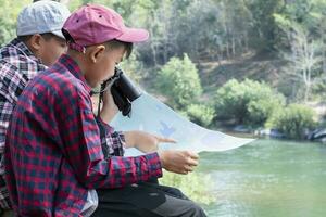 Asian boys holding binoculars and national map and reading map details about bird watching and fish watching by the river bank in local national park, nature study and environment learning concept. photo