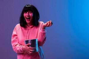 Cheerful excited awesome brunet woman in pink hoodie with popcorn hold hand up open mouth posing isolated in blue violet color light studio background. Neon party Cyberpunk Cinema concept. Copy space photo