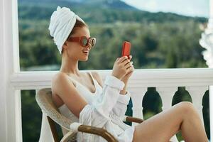 hermosa mujer disfrutando el Mañana en el balcón mirando a el móvil teléfono pantalla montaña ver foto