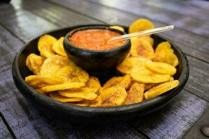 plátano papas fritas servido con Colombiana tradicional hogao en parte superior de un de madera mesa foto