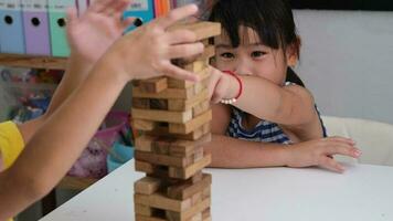 söt asiatisk syskon har roligt spelar jenga tillsammans. två barn spelar jenga styrelse spel på tabell i rum på Hem. trä- pussel är spel den där öka intelligens för barn. video