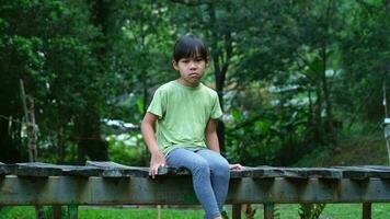 Cute little girl upset sitting on a wooden bridge near the river in summer. little girl sitting alone on a wooden bridge looking at the river outdoors. video