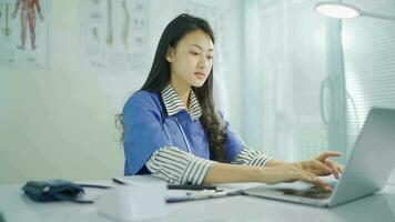 Young female doctor in white coat working on laptop in modern hospital office room, therapist typing on computer consult patient online, makes research, noting, takes useful information from internet video