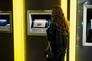 Back of young brunette girl withdrawing money from credit card at yellow ATM. photo