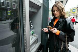 joven mujer en cuero chaqueta insertando un crédito tarjeta a Cajero automático exterior, mientras mirando en teléfono inteligente foto