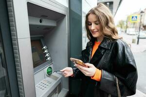 joven mujer en cuero chaqueta insertando un crédito tarjeta a Cajero automático exterior, mientras mirando en teléfono inteligente foto