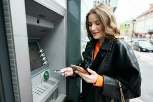 Young woman in leather jacket inserting a credit card to ATM outdoor, with surprised face while looking on smartphone. photo