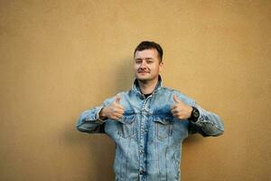 Portrait of stylish man in jeans jacket against yellow wall show thumbs up. photo