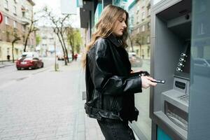 joven mujer en cuero chaqueta insertando un crédito tarjeta a Cajero automático exterior. foto