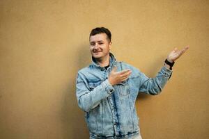 Portrait of stylish man in jeans jacket against yellow wall hand gesture at copy space. photo