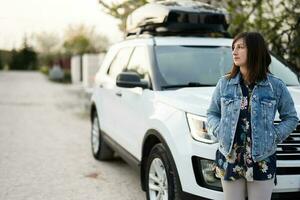 Stylish woman in jeans jacket standing near white american suv car with roof rack box. photo