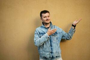 Portrait of stylish man in jeans jacket against yellow wall hand gesture at copy space. photo