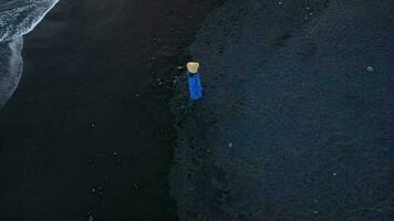 parte superior ver de un niña en un azul vestir y sombrero caminando en el playa con negro arena, espumoso olas de el atlántico océano. tenerife, canario islas, España video