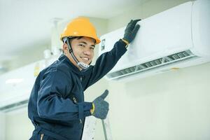 un asiático joven técnico Servicio hombre vistiendo azul uniforme comprobando, profesional aire acondicionador instalador mantener moderno interior aire acondicionador espacio para texto foto