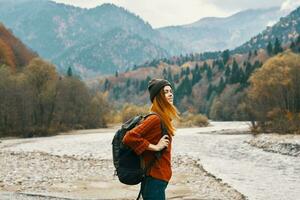 alegre mujer turista con un mochila mira a el montañas cerca el río en el banco foto