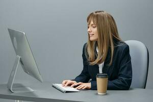 Millennial blonde businesswoman worker in blue jacket using desktop computer with takeaway coffee sitting at workplace in gray office. Remote Job, Technology And Career Profession Concept. Copy space photo