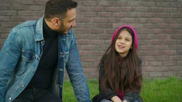 Dad and daughter are sitting and talking against the backdrop of grass and a brick wall. Concept of support and happy video