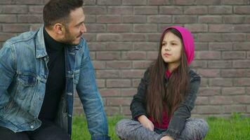 Dad and daughter are sitting and talking against the backdrop of grass and a brick wall. Concept of mutual understanding, support and happy family video