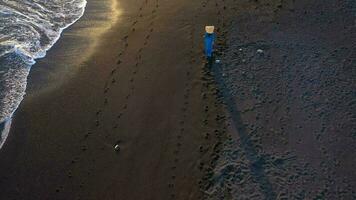 Top view of a girl in a blue dress and hat walking on the beach with black sand, foaming waves of the Atlantic Ocean. Tenerife, Canary Islands, Spain video