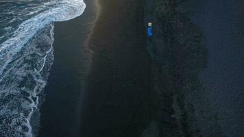 Top view of a girl in a blue dress and hat walking on the beach with black sand, foaming waves of the Atlantic Ocean. Tenerife, Canary Islands, Spain video