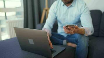 Mens zittend Aan een knus sofa en werken Aan laptop en smartphone. concept van afgelegen werk. huis kantoor video