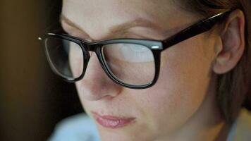 Woman in glasses looking on the monitor and surfing Internet at night. The monitor screen is reflected in the glasses. Extremely close up. Work at night. Home Office. Remote work video