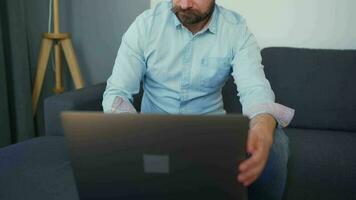 Mann Sitzung auf ein gemütlich Sofa und Arbeiten auf Laptop und Smartphone. Konzept von Fernbedienung Arbeit video