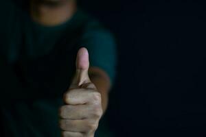 young man showing gesture thumbs up or giving approval on dark black background. selective focus on finger photo