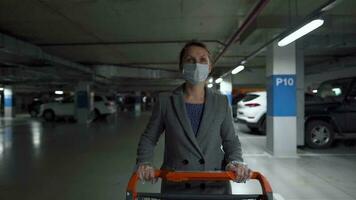vrouw in een medisch masker en beschermend handschoenen wandelingen met een kruidenier kar door een ondergronds parkeren kavel. aankoop gedurende covid-19 coronavirus pademie video
