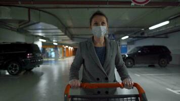 Woman in a medical mask and protective gloves walks with a grocery cart through an underground parking lot. Purchase during Covid-19 coronavirus pademia video