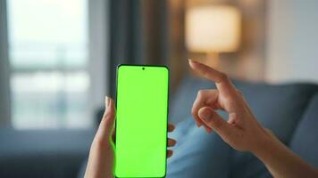 Woman at home lying on a couch and using smartphone with green mock-up screen in horizontal landscape mode. Girl browsing Internet, watching content, videos, blogs. POV. video