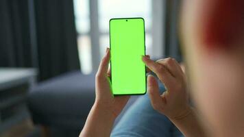 Woman at home lying on a couch and using smartphone with green mock-up screen in horizontal landscape mode. Girl browsing Internet, watching content, videos, blogs. POV. video