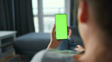 Woman at home lying on a couch and using smartphone with green mock-up screen in horizontal landscape mode. Girl browsing Internet, watching content, videos, blogs. POV. video