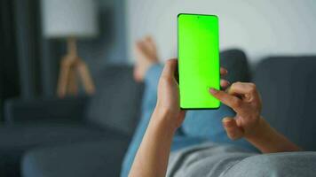 Woman at home lying on a couch and using smartphone with green mock-up screen in horizontal landscape mode. Girl browsing Internet, watching content, videos, blogs. POV. video