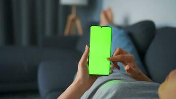 Woman at home lying on a couch and using smartphone with green mock-up screen in horizontal landscape mode. Girl browsing Internet, watching content, videos, blogs. POV. video