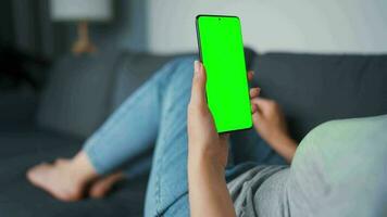 Woman at home lying on a couch and using smartphone with green mock-up screen in horizontal landscape mode. Girl browsing Internet, watching content, videos, blogs. POV. video