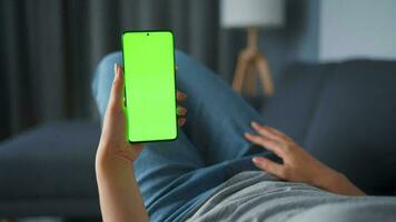 Woman at home lying on a couch and using smartphone with green mock-up screen in horizontal landscape mode. Girl browsing Internet, watching content, videos, blogs. POV. video