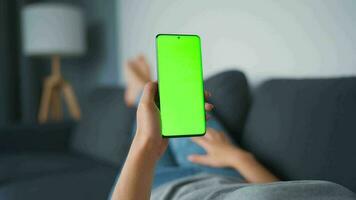 Woman at home lying on a couch and using smartphone with green mock-up screen in horizontal landscape mode. Girl browsing Internet, watching content, videos, blogs. POV. video